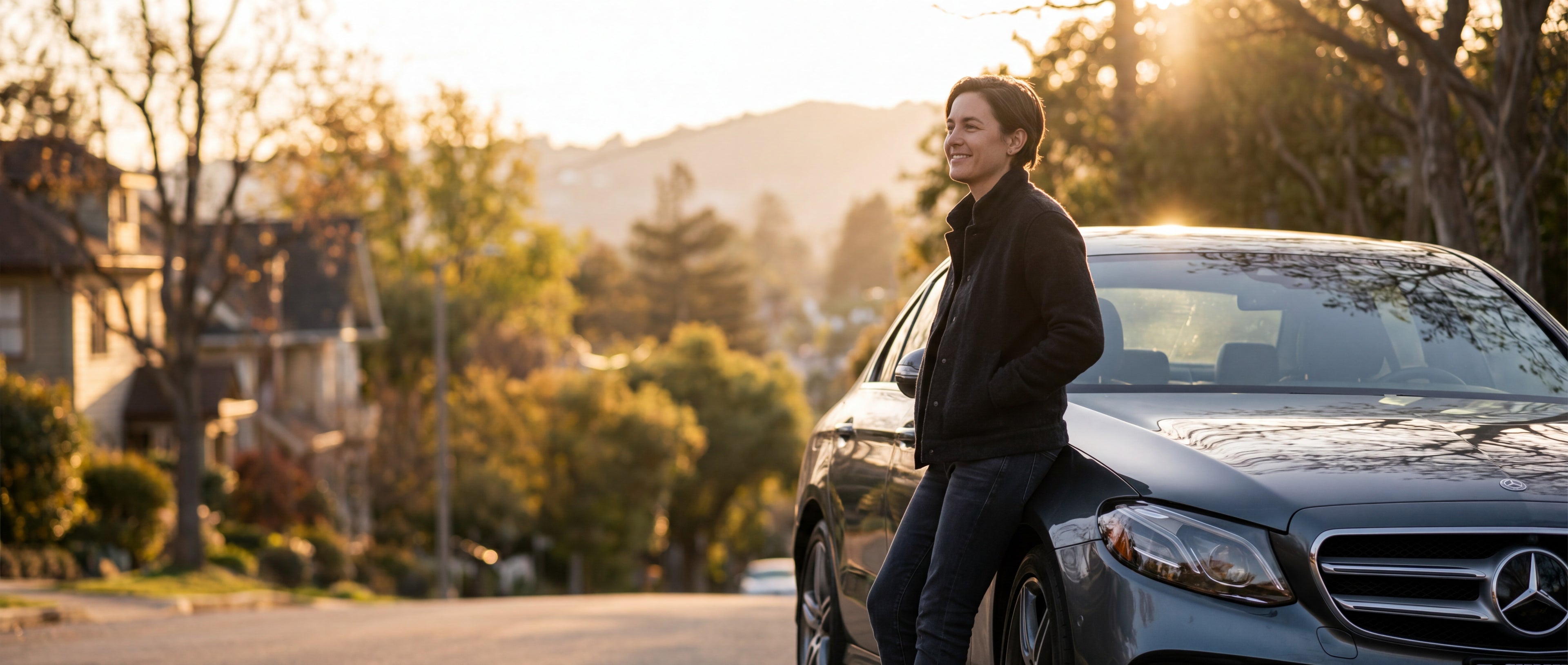 A man in a black jacket and jeans stands next to a dark gray Mercedes-Benz sedan on a sunlit suburban street.