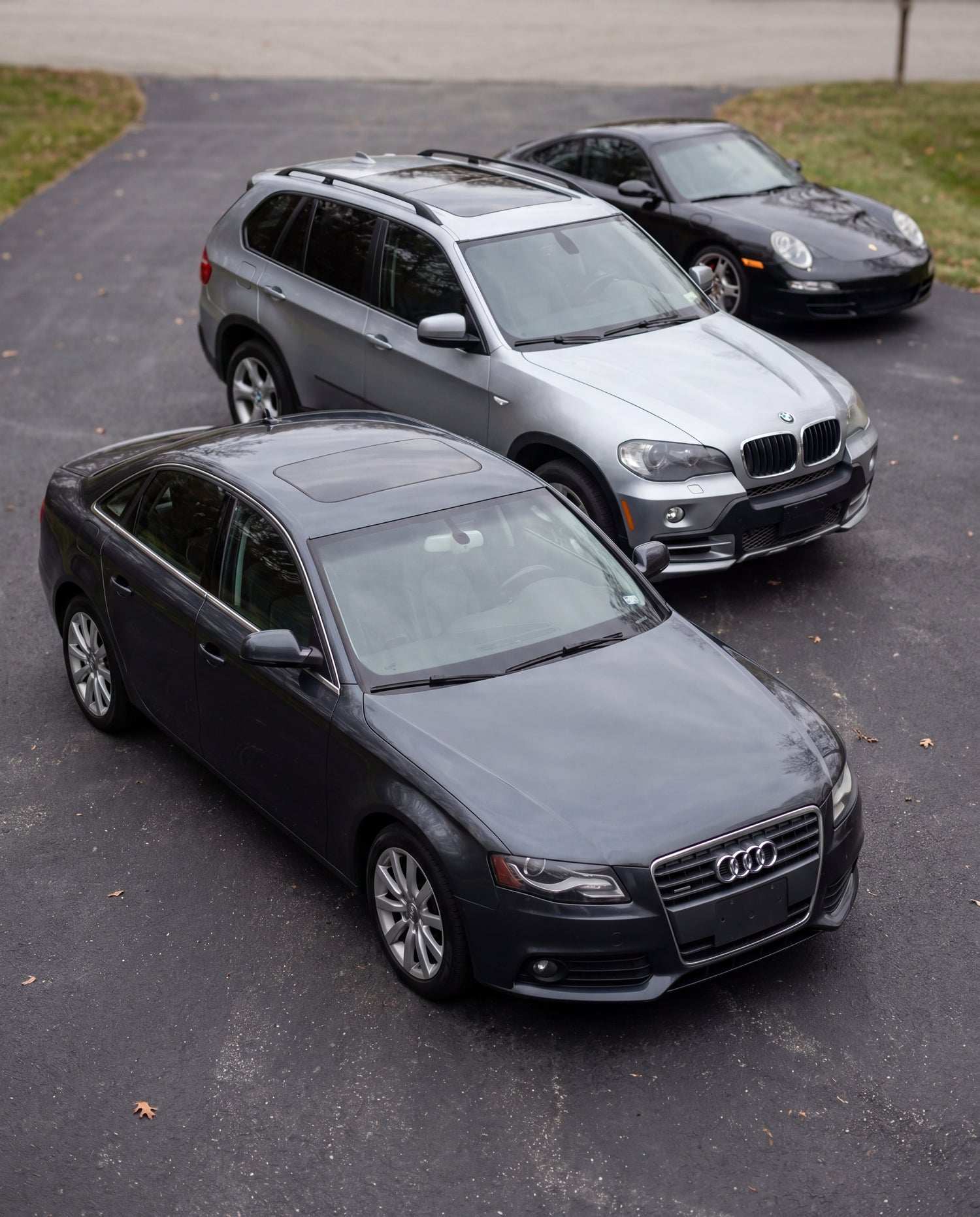 Three cars parked on an asphalt surface: a dark gray Audi sedan in the foreground, a silver BMW X5 SUV behind it, and a black Porsche behind the BMW.